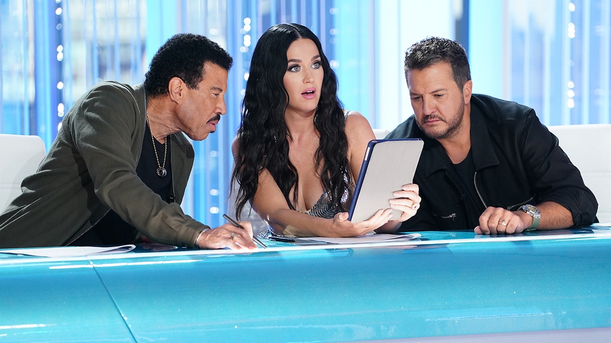 Lionel Richie leans over and looks at Katy Perry holding something in the middle as Luke Bryan leans over from the far right during "American Idol" auditions