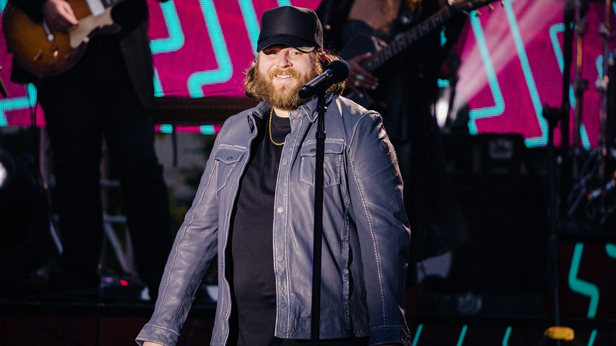 Nate Smith in a black hat and blue jacket performs on stage from the CMT Music Awards