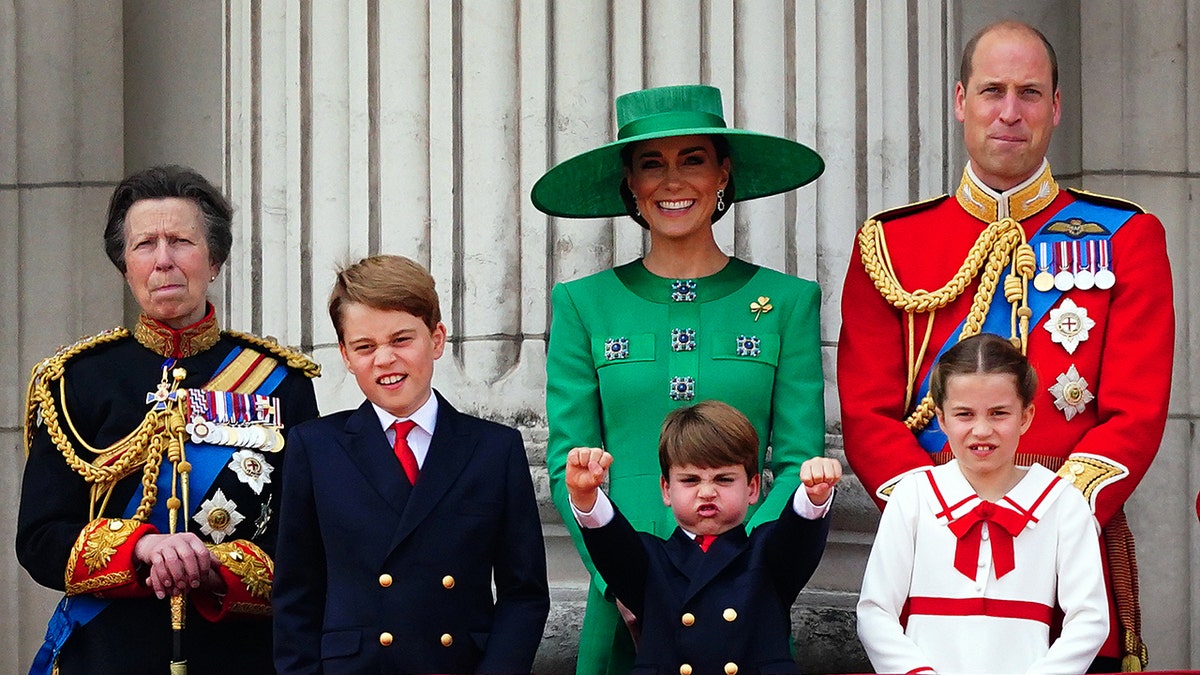 The Princess Royal stands beside Prince George, who display his own funny face, next to Prince Louis outstretching his arms and showing his fists, with Kate Middleton laughing behind her son, and Prince William trying to hold in a laugh, in front of Princess Charlotte