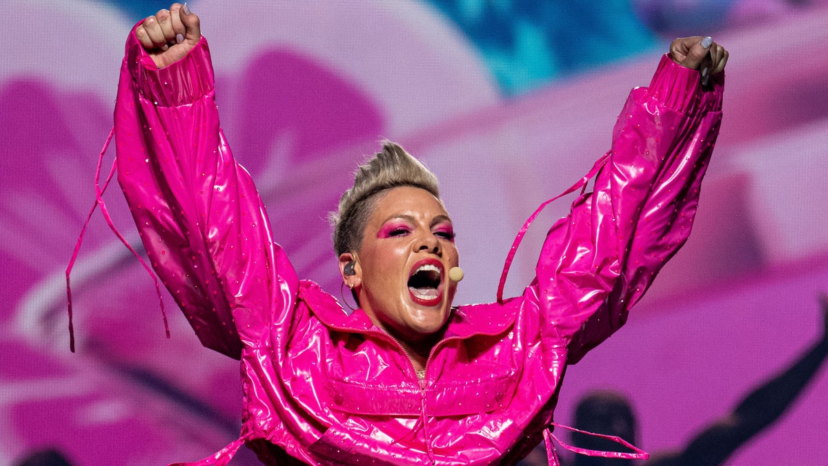 Pink throws her arms up in the air wearing a hot pink latex-like jacket while on stage