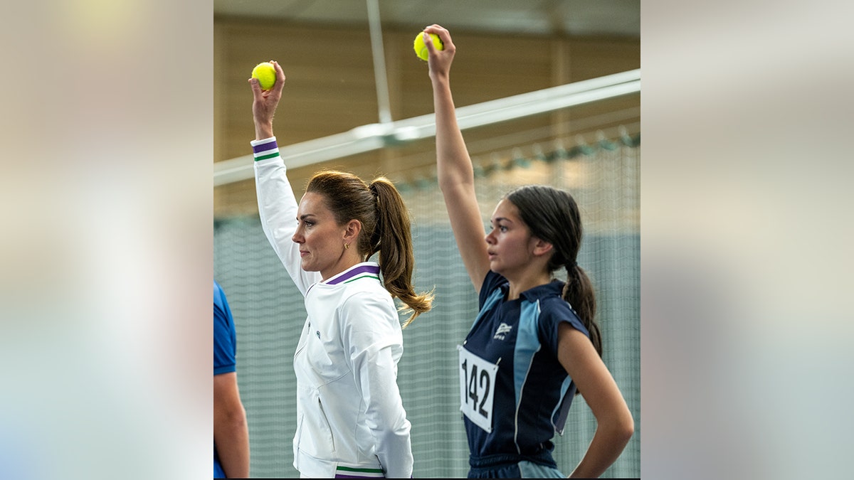 Kate Middleton holds a ball up in the air as if she were to feed it to the player on the baseline next to another young girl