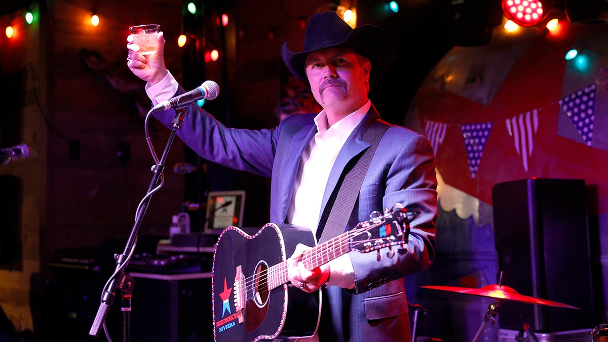 John Rich raises a glass on stage with a guitar swung across his chest in Nashville