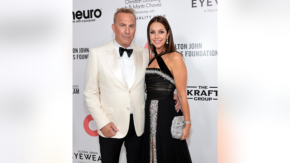 Kevin Costner soft smiles in a white tuxedo with his wife Christine in a black criss-cross gown at the Elton John AIDS Foundation party