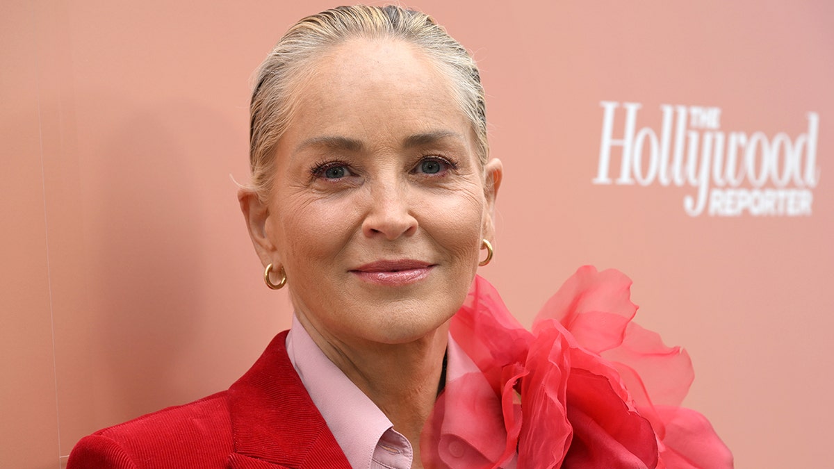 Sharon Stone soft smiles on the red carpet at The Hollywood Reporter Raising Our Voices DEIA Luncheon wearing a red suit with a large flower on the shoulder