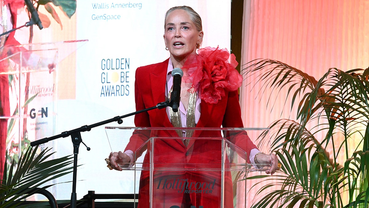 Sharon Stone holds the glass podium as she speaks to the "Raising Our Voices" luncheon crowd wearing a red suit with a flower