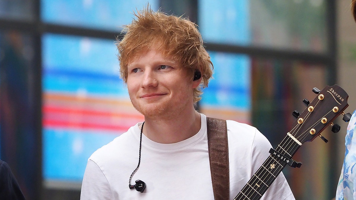 Ed Sheeran smiles wearing a white shirt in NYC with his guitar strapped over his arm and earpiece hanging on his shirt