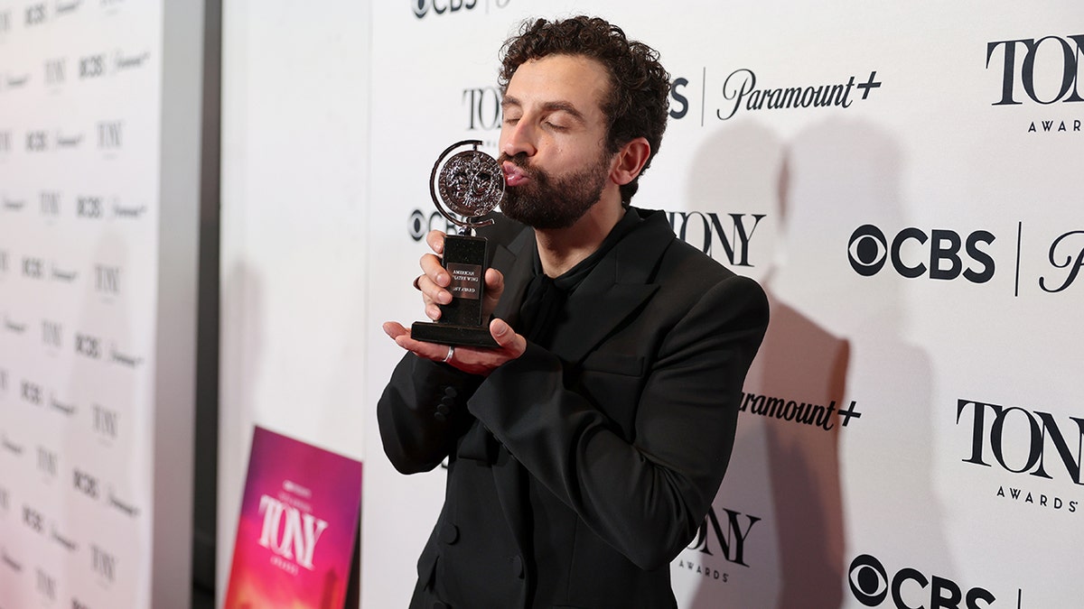 Brandon Uranowitz in a black jacket kisses his Tony Award