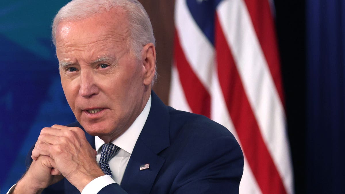 President Joe Biden holds his hands together as he sits in front of an American flag