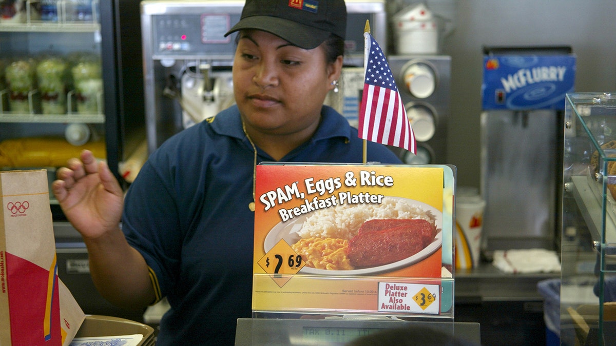 Seorang kasir di lokasi McDonald's di Wahiawa, Hawaii, berdiri di belakang iklan SPAM, Eggs and Rice Breakfast Platter yang baru pada bulan Juni 2002. Penduduk Hawaii mengonsumsi lebih banyak SPAM dibandingkan dengan penduduk negara bagian AS lainnya.