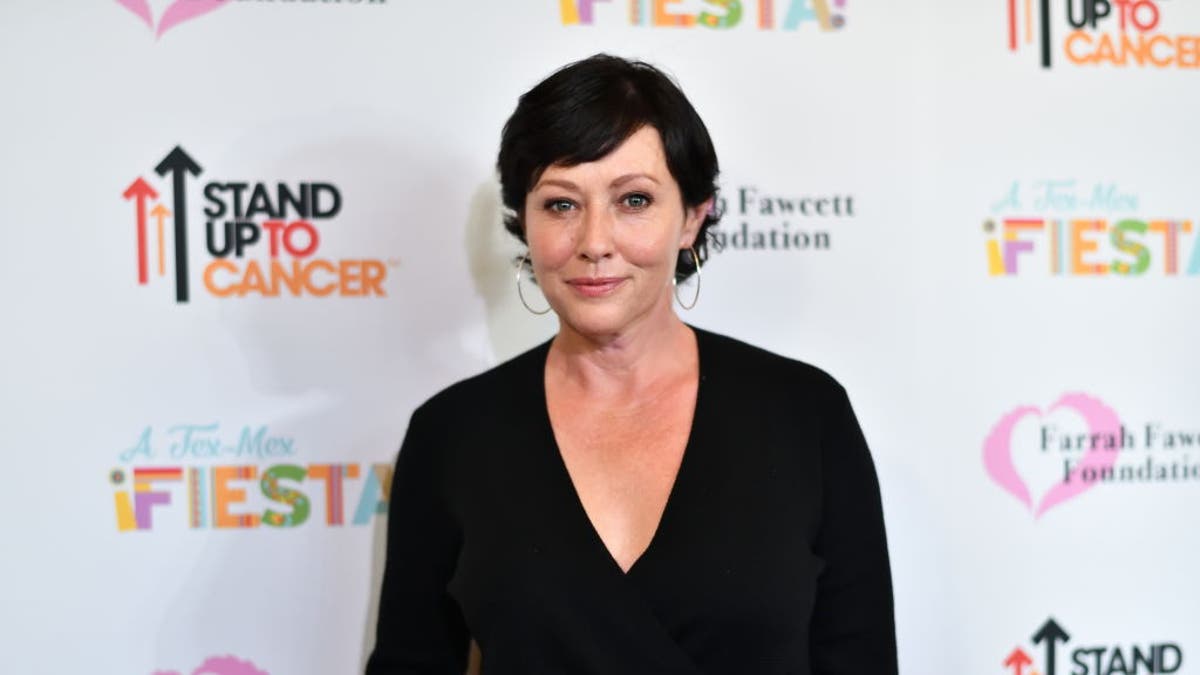 Shannen Doherty in a black v-neck dress with short black hair on the carpet