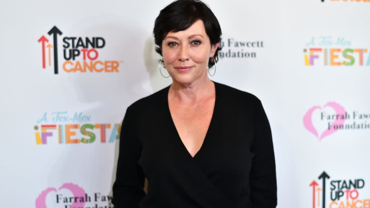 Shannen Doherty with short black hair and a black blouse on the carpet