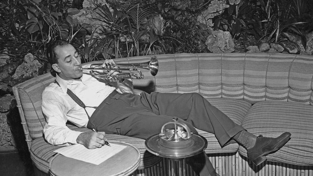 Louis Prima lounging on the couch while playing his trumpet