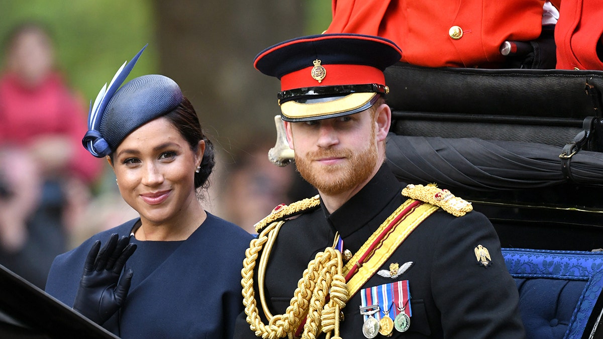 Meghan Markle in a blue dress and matching hat sitting next to Prince Harry in his military uniform