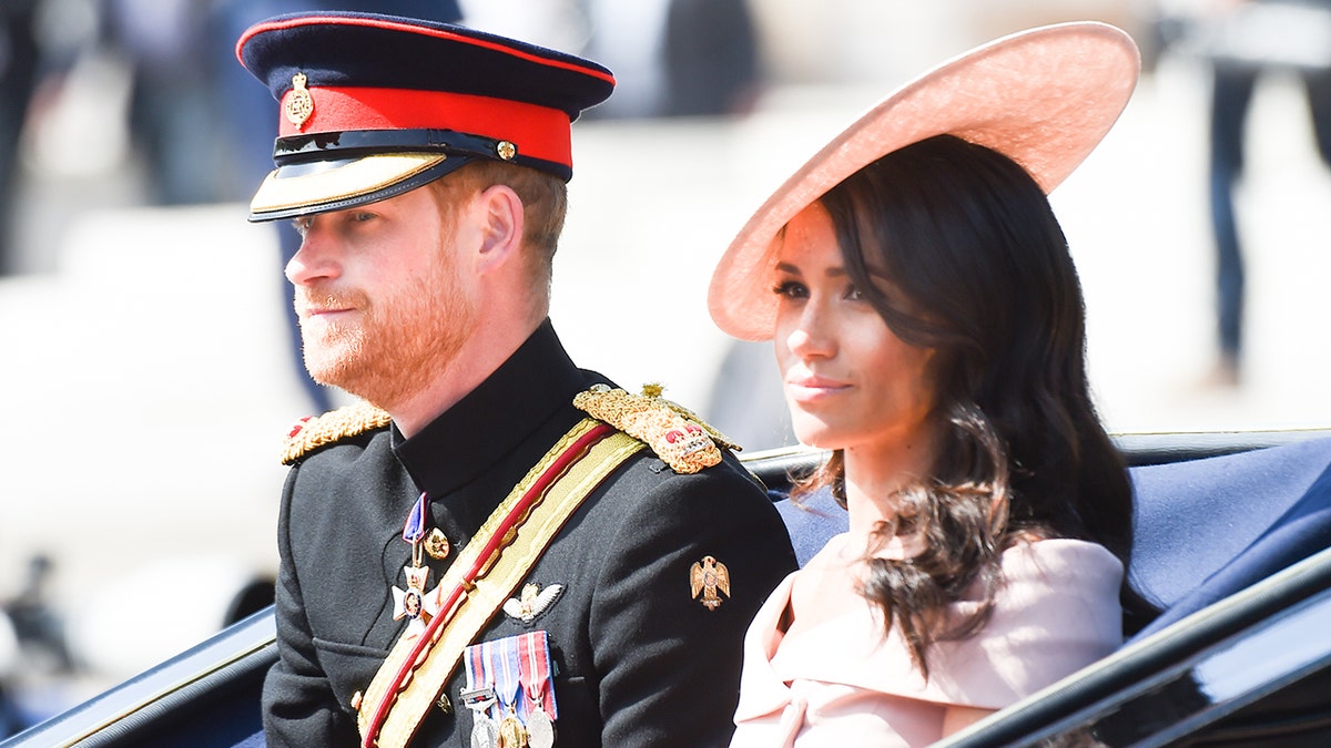 A close-up of Meghan Markle wearing a pink dress and matching hat sitting next to Prince Harry in his military uniform