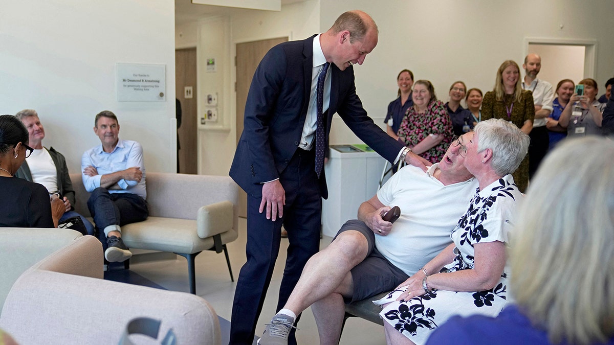 Prince William in a blue suit laughing with a man wearing a white shirt and shorts at a cancer centre