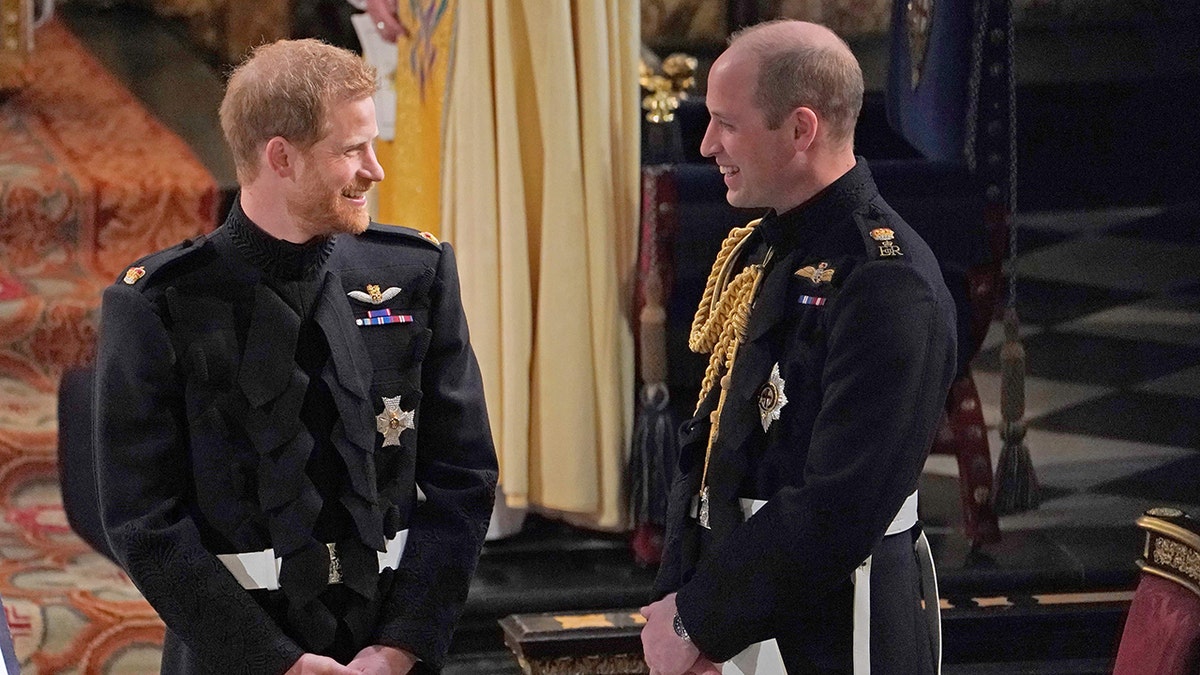 Prince William and Prince Harry smiling at each other wearing suits with medals