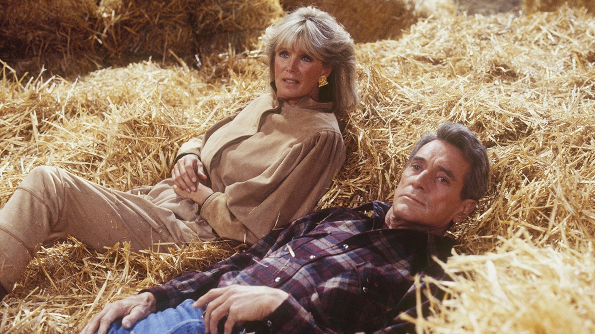 Linda Evans wearing a tan dress laying on hay with Rock Hudson