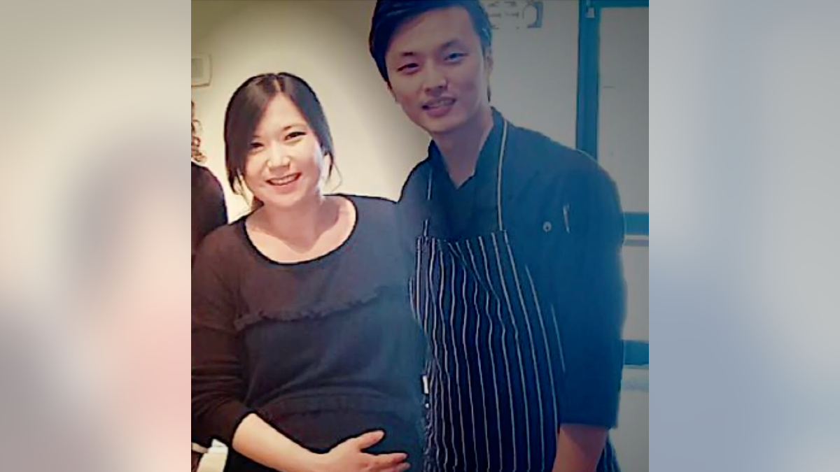 Eina Kwon holds her belly as she stands smiling next to her husbandd