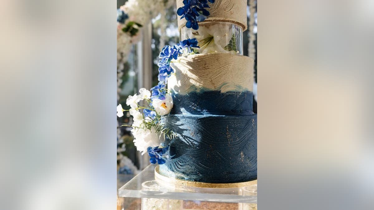 Wedding cake with a base tier that has swirling blue icing that eventually switches to white is on display. The cake has decorative gold flecks and blue and white flowers.