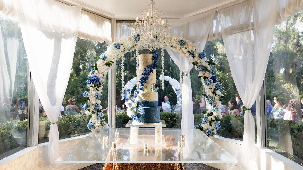 A five-tier wedding cake with a blue base that fades into white. There's an acrylic cake separator in the middle. The cake is set up inside an indoor ballroom and sits in front of a flower arch.