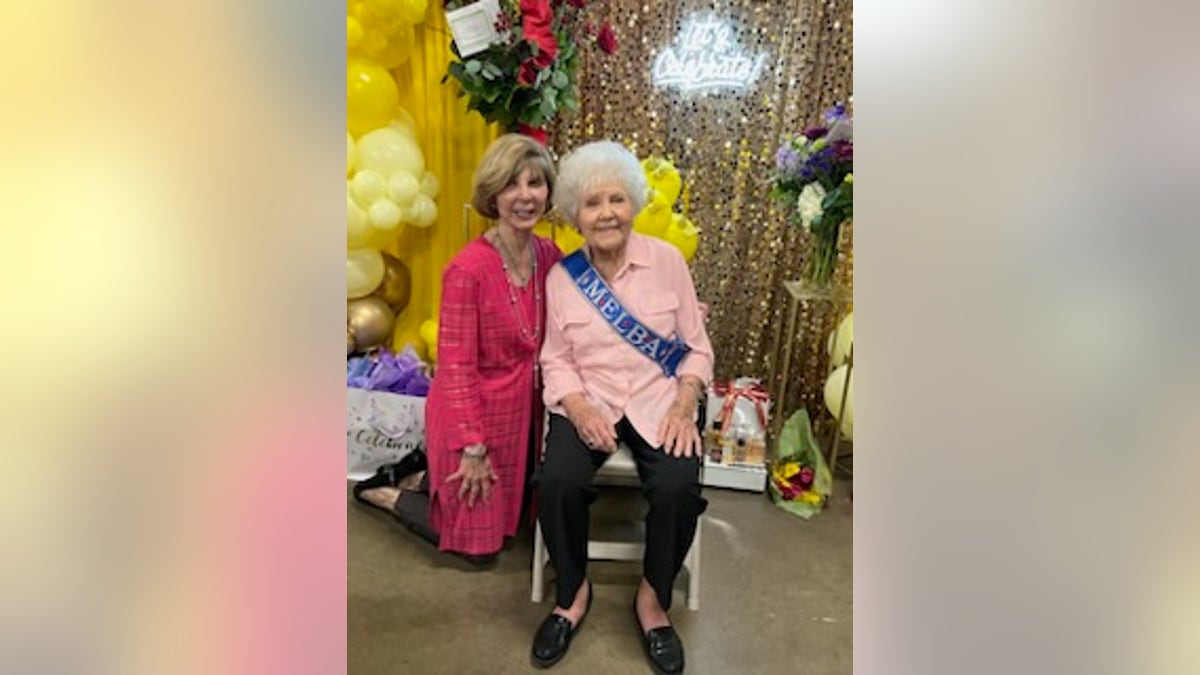 Melba wearing a pink shirt is sitting next to drew dillar who is wearing a pink cardigan, they are both in front of a gold curtain and yellow baloons.