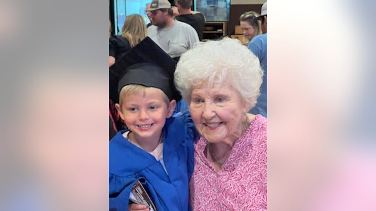 Young boy on the left held up by his grandmother on the right, the boy is wearing a graduation gown