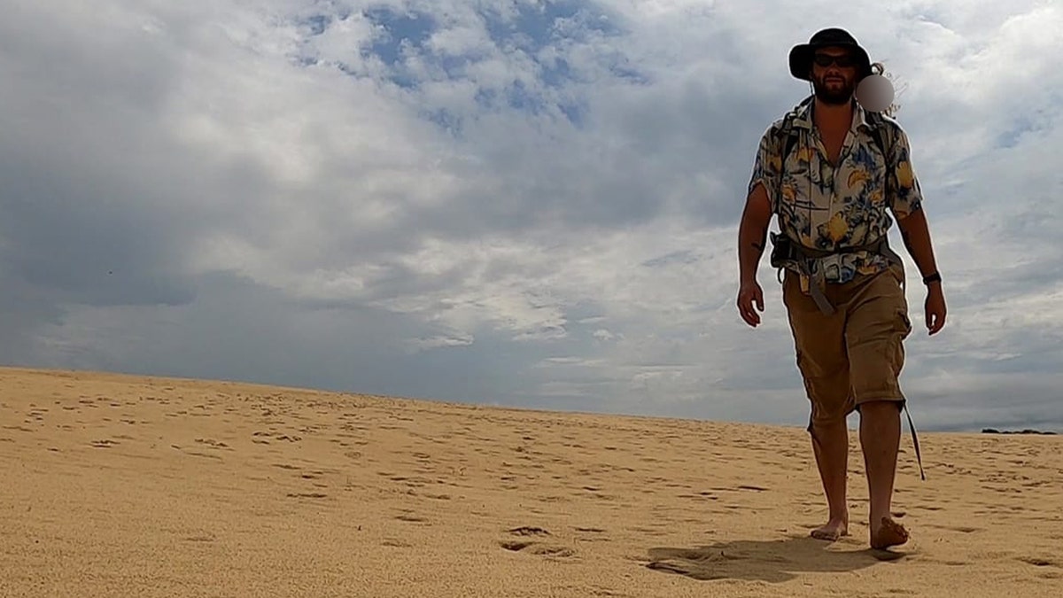A man in khaki shorts, hawaiian shirt, sunglasses and wide-brim hat walking barefoot in the sand under a cloudy sky