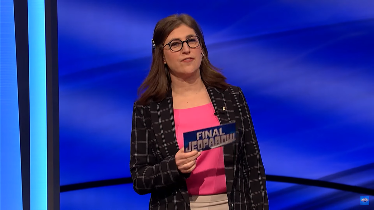 Mayim Bialik in a hot pink top and checkered blazer holding a card on "Jeopardy!"