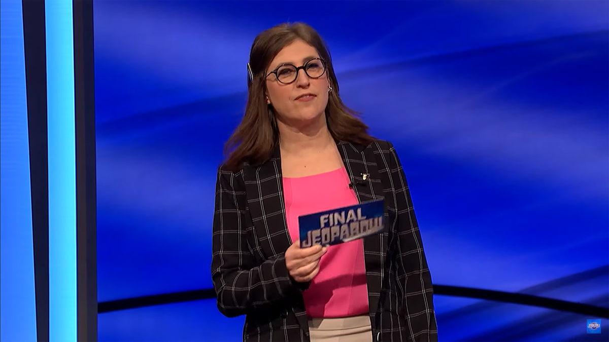 Mayim Bialik con un top rosa acceso e un blazer a scacchi con in mano una carta "Pericolo!"