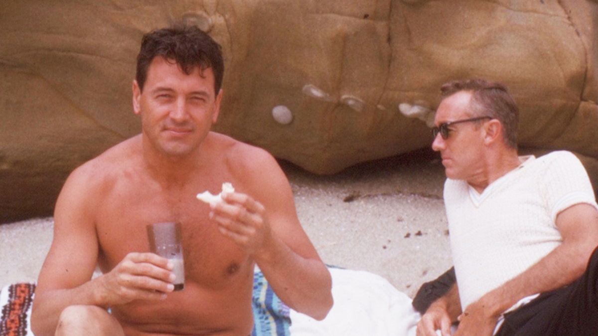 Rock Hudson shirtless on the beach next to a male friend wearing a white shirt and sunglasses