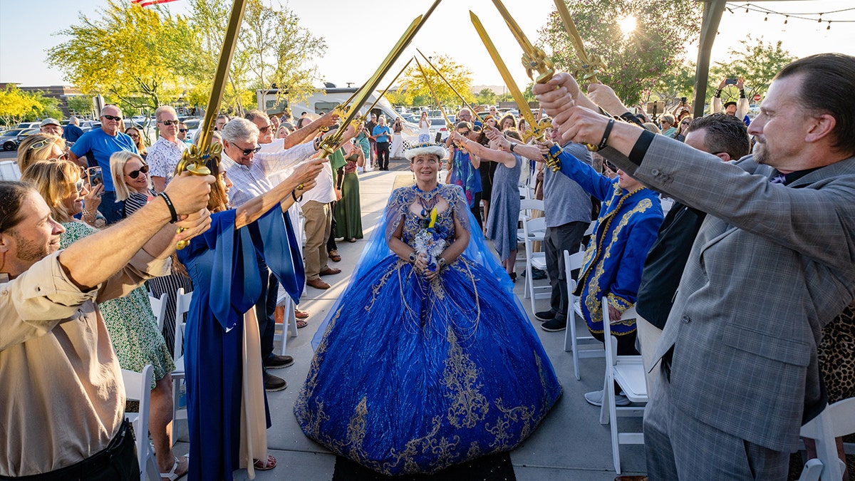 Jamie West walks down the aisle in a blue and gold wedding gown and crown while guests hold up decorative swords.