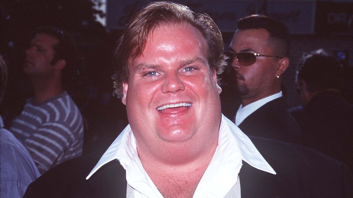 Chris Farley in a suit jacket and a white collared shirt smiles 