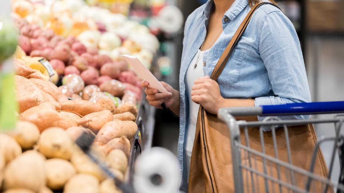 Woman holds shopping list and purse while she stands in front of potato bins at a supermarket.