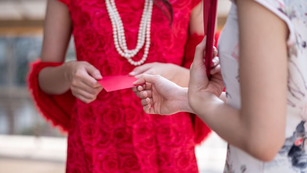 Woman in red dress and pearl necklace hands a red envelope to another woman in a floral white dress.