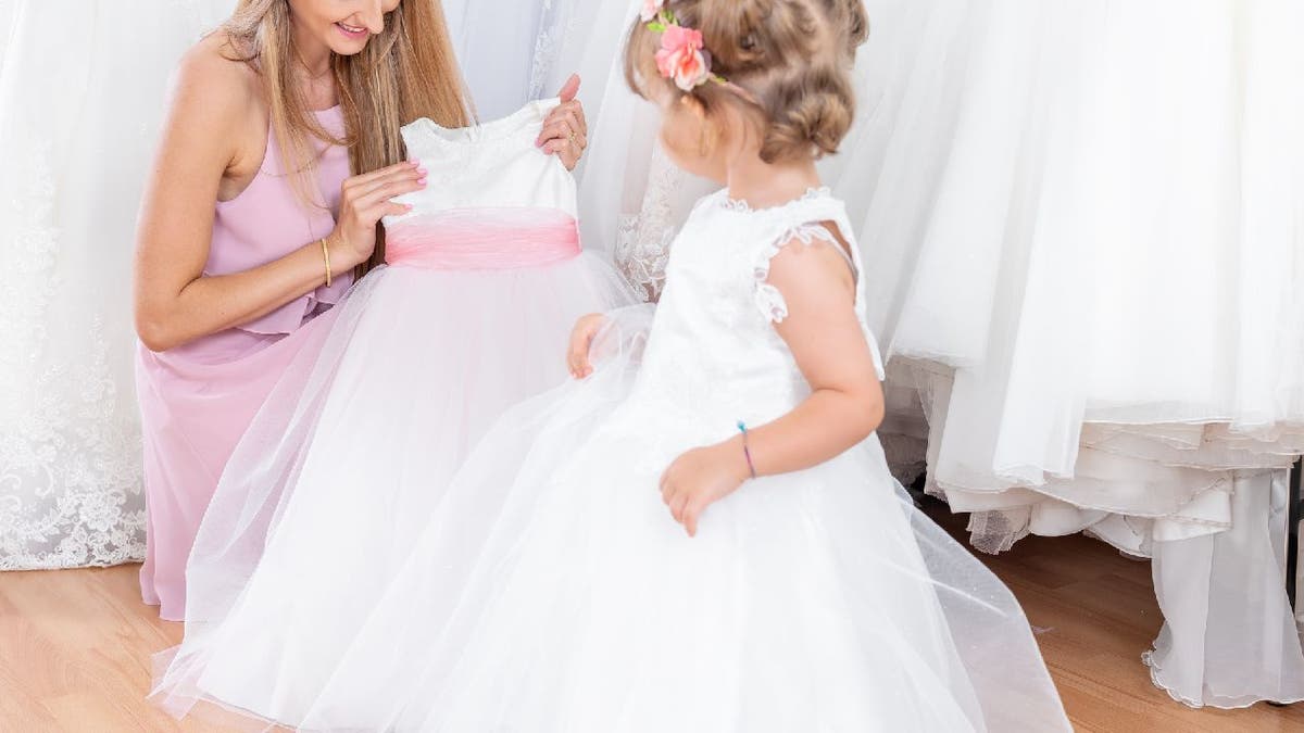 Woman holds up a flower girl dress in front of a young girl.