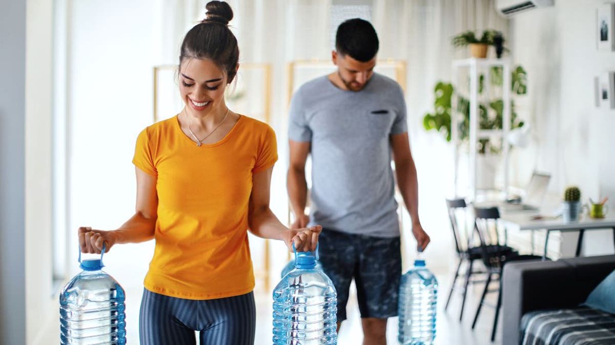 A man and woman carry gallon water bottles while wearing workout clothes.