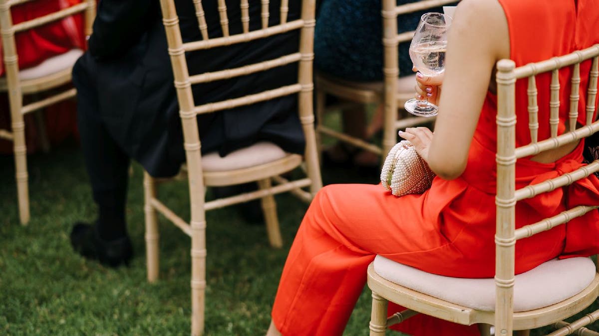 Woman wears a red jumpsuit while sitting on a lawn chair during what appears to be a wedding ceremony. Another woman a short distance can be seen wearing red as well.