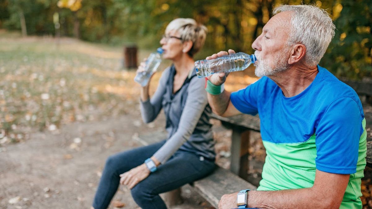 An older man and woman stop to drink water while sitting on a park bench.