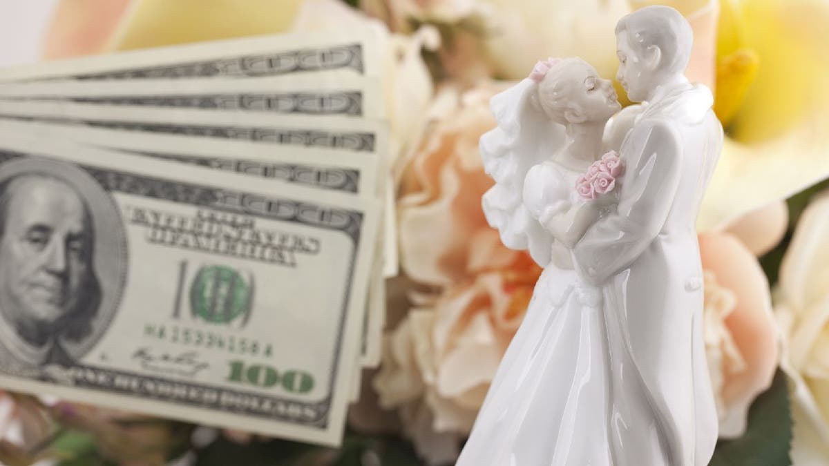 Hundred dollar bills next to bride and groom cake topper and bouquet.