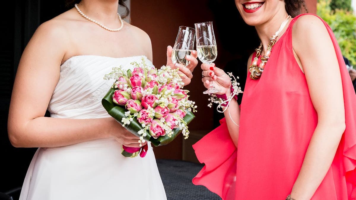 A bride wears a achromatic dress and holds a bouquet while she toasts champagne pinch a wedding impermanent who's wearing a reddish dress.