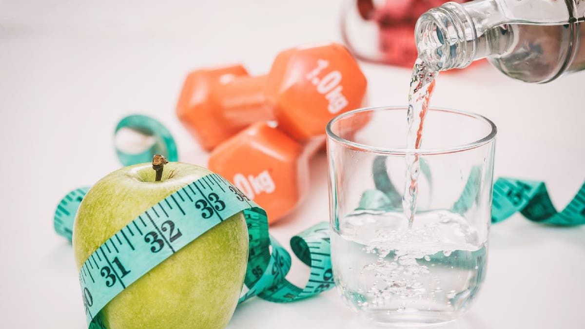 Glass of water gets filled next to green apple, dumbbells and measuring tape.