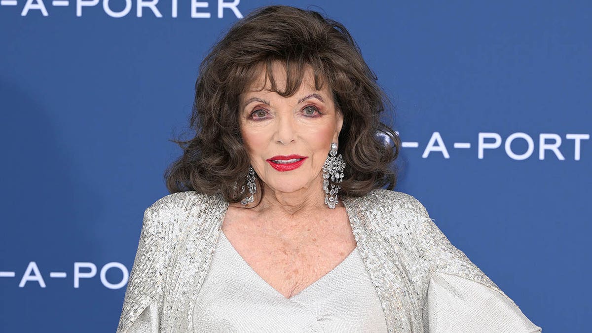 Joan Collins stands with her hand on hip on red carpet