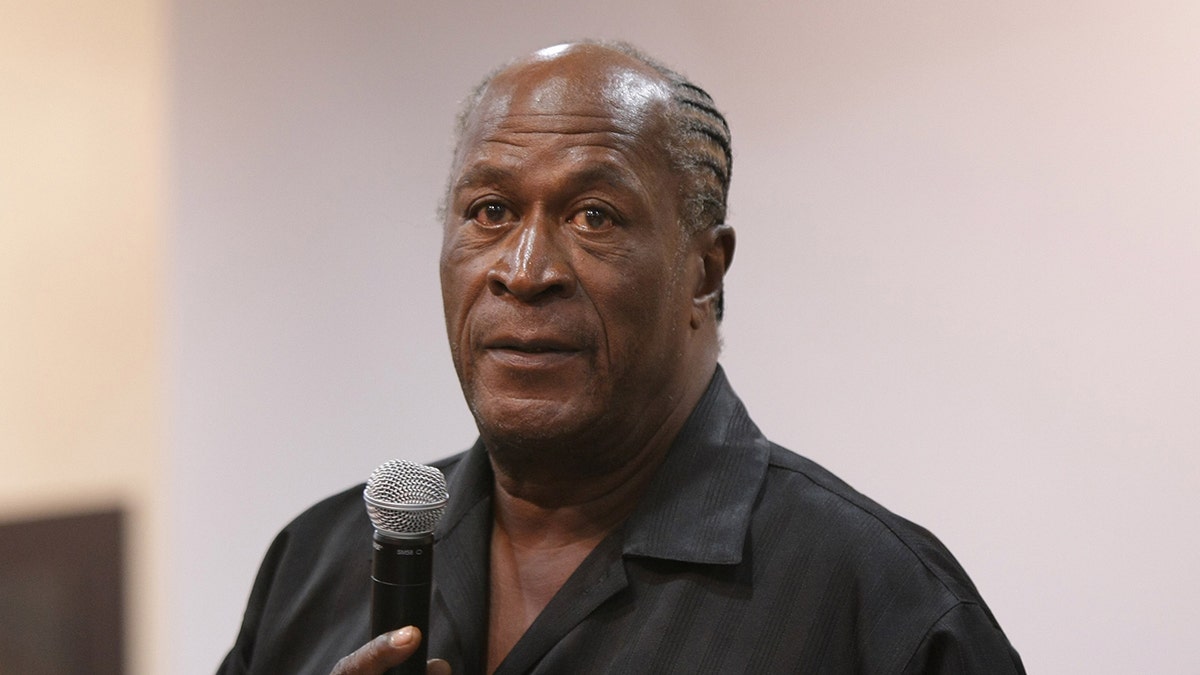 John Amos holds a microphone while speaking to a crowd