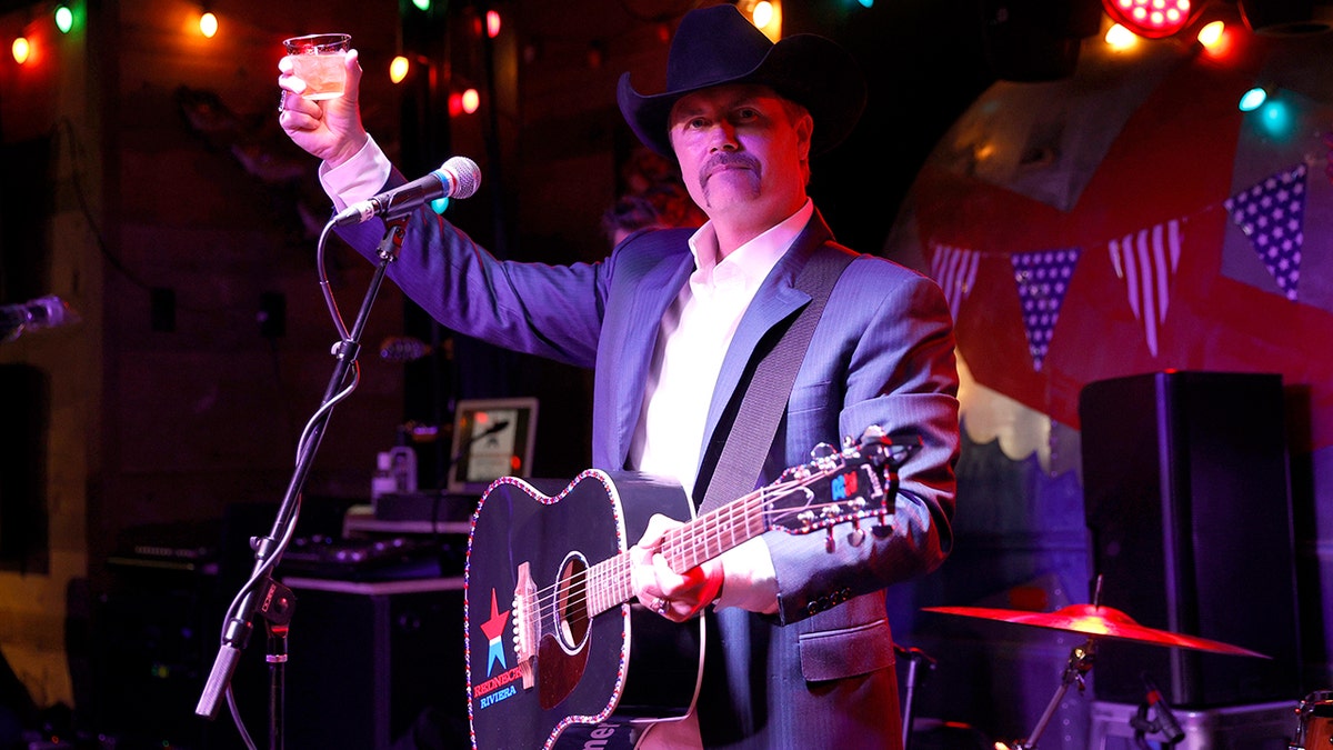 John Rich holds whiskey glass in the air with guitar strung around his neck at Nashville bar