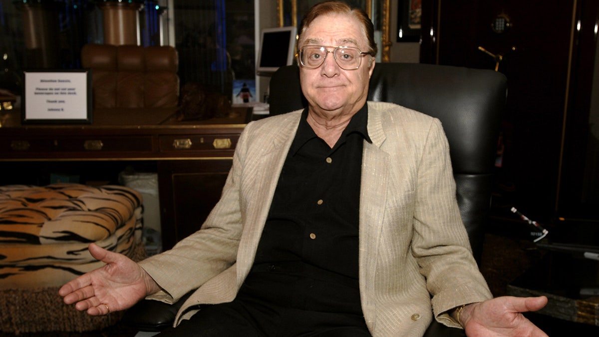 Pat Cooper sits on a leather chair while wearing a khaki blazer