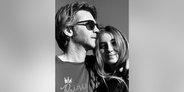 A close-up black and white photo of Prince Emanuele Filiberto wearing sunglasses and a dark shirt kissing his daughters forehead