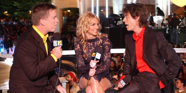 Kurt Loder interview Britney Spears in a black dress who looks at Mick Jagger in a red shirt and black suit