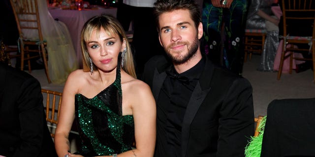 Miley Cyrus in a green and black gown sits at a table next to Liam Hemsworth in all black at The Met