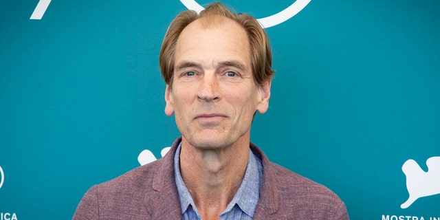 Julian Sands soft smiles on the carpet in a light burgundy jacket and blue shirt