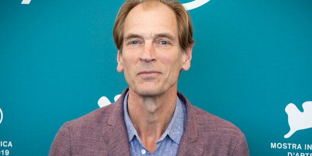 Julian Sands soft smiles on the carpet in a light burgundy jacket and blue shirt